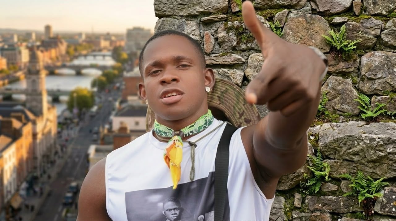 SECKY pointing toward the camera in front of a stone wall, with a city riverside skyline behind him